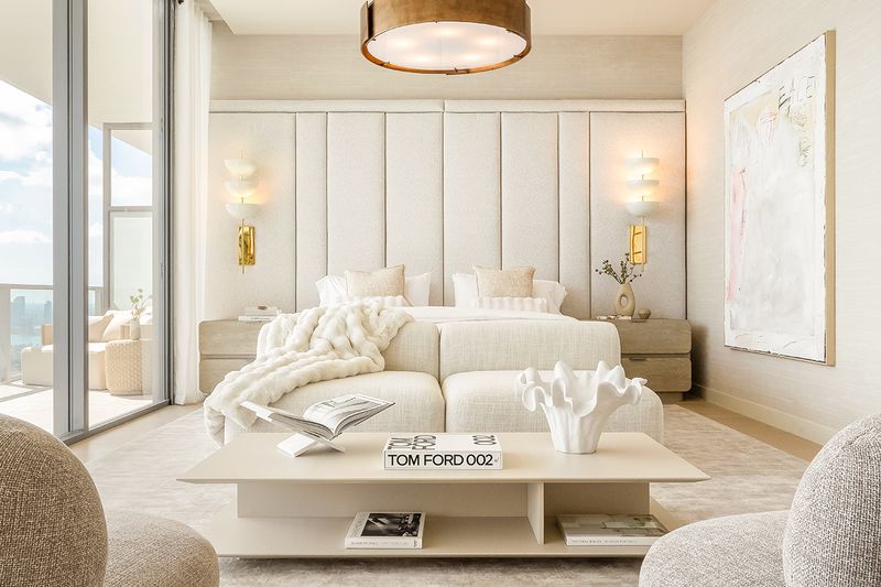 An all-white bedroom with an oversized upholstered headboard, layered bed, loveseat, coffee table, and a furnished balcony.