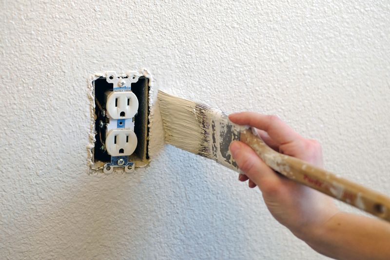 Anonymous person using a bristled paint brush to paint around an electrical outlet with the cover removed.