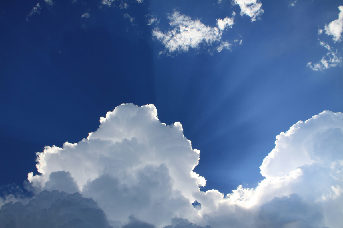 A picture of white, puffy clouds in a bright blue sky with the sun peeking through.