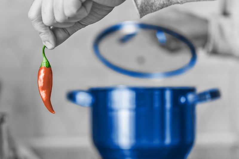 Hand holding chili pepper and lifting the lid off a pot